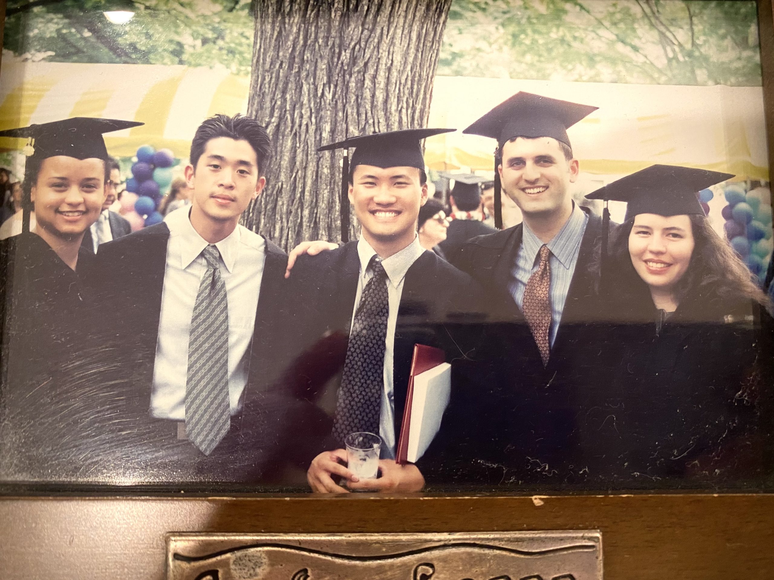 Five graduates in caps and gowns standing together outdoors, smiling and holding diplomas under a tree.