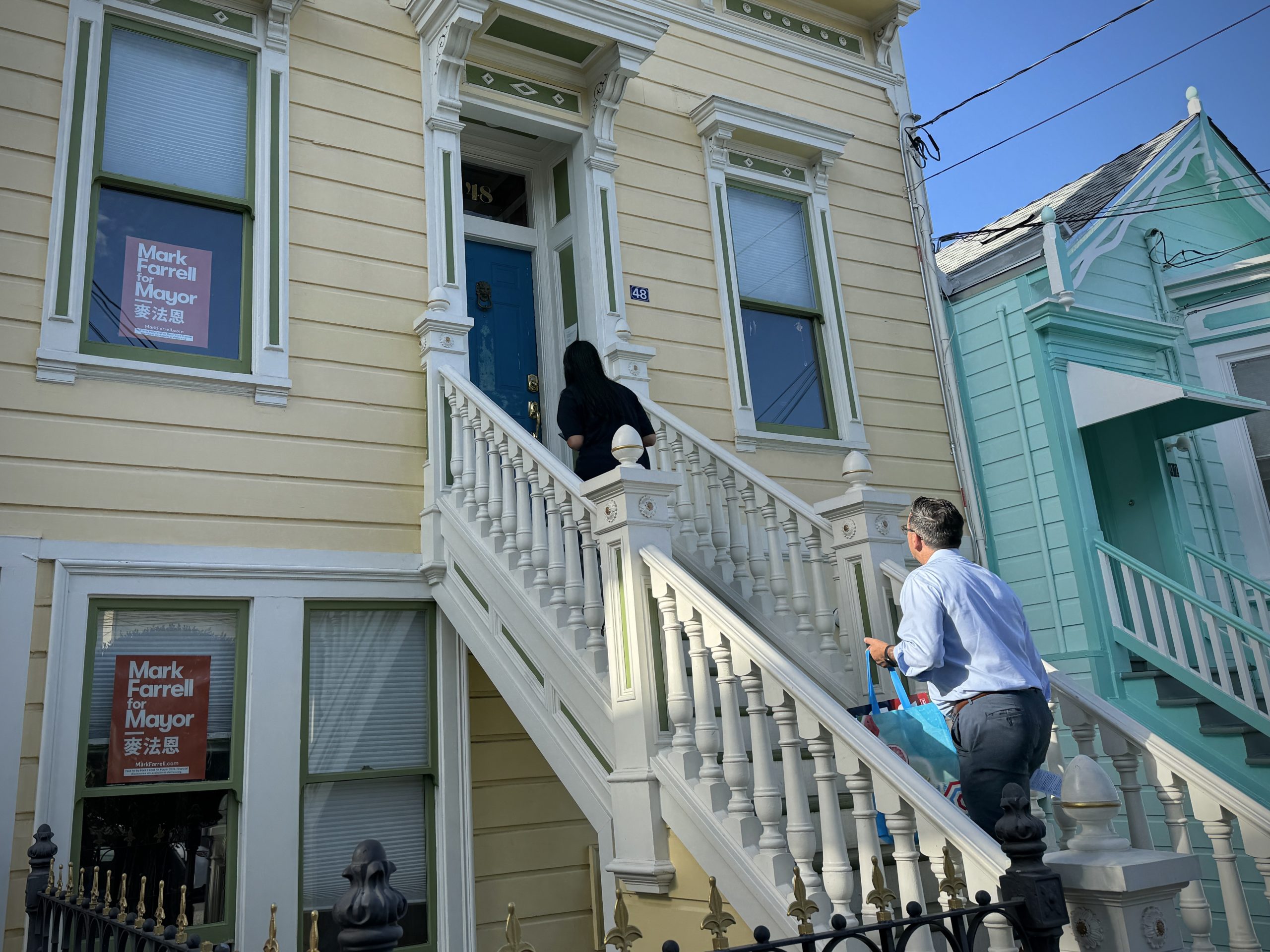 Two people approach the front steps of a beige house with "Mark Farrell for Mayor" signs in the windows.