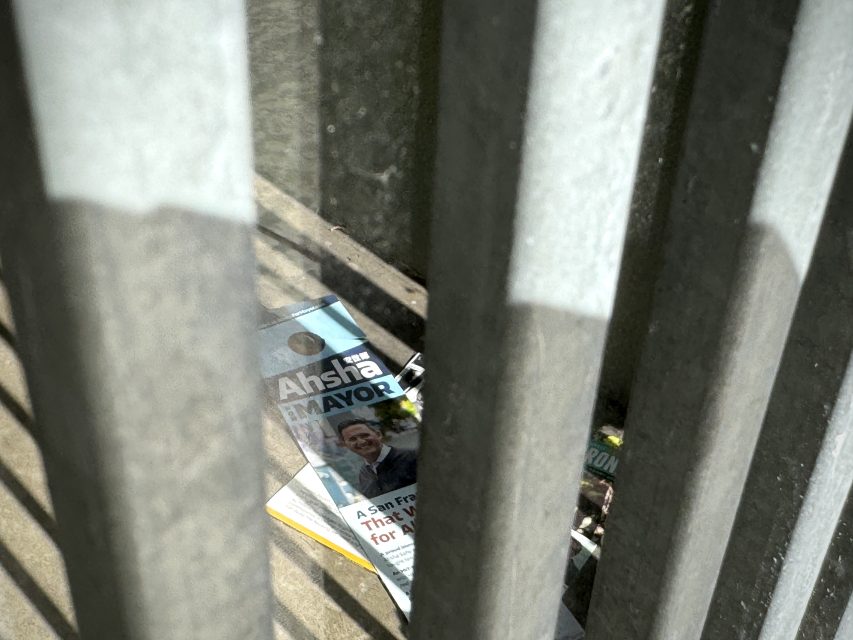 A mayoral campaign flyer featuring a smiling candidate lies discarded on the ground behind metal bars.
