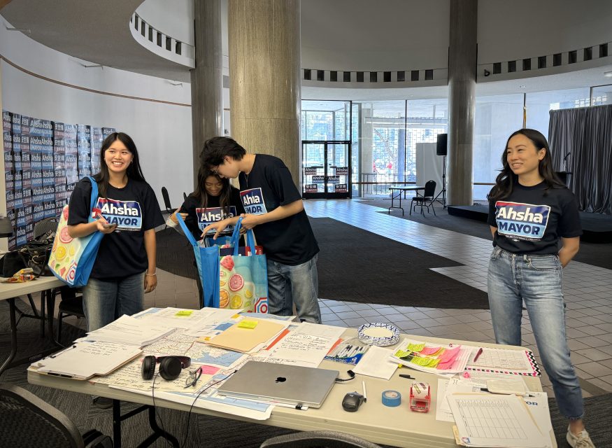 Four individuals in "Ahsha for Mayor" T-shirts stand by a table with office supplies, campaign materials, and bags. They appear to be in a campaign office with posters on the walls.