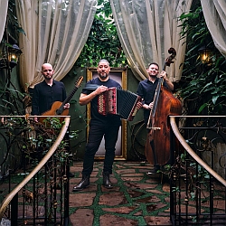 Three men stand in a garden setting, with the central figure playing an accordion, and the men on the left and right holding a guitar and double bass, respectively.