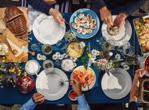 Overhead view of a table with people eating various dishes, including bread, deviled eggs, salad, and beverages. The table is set with plates, cutlery, a cutting board, and floral arrangements.