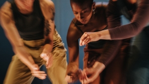 Three individuals, slightly blurred in motion, perform a dance with arms extended forwards against a neutral backdrop.
