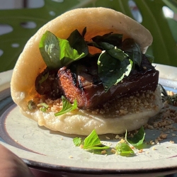 A steamed bao bun filled with grilled meat, fresh greens, and crushed peanuts is placed on a plate with a simplistic design.