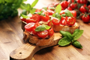 Slices of toasted bread topped with cherry tomatoes, basil leaves, and cheese, placed on a wooden cutting board. Fresh basil and tomatoes are in the background.