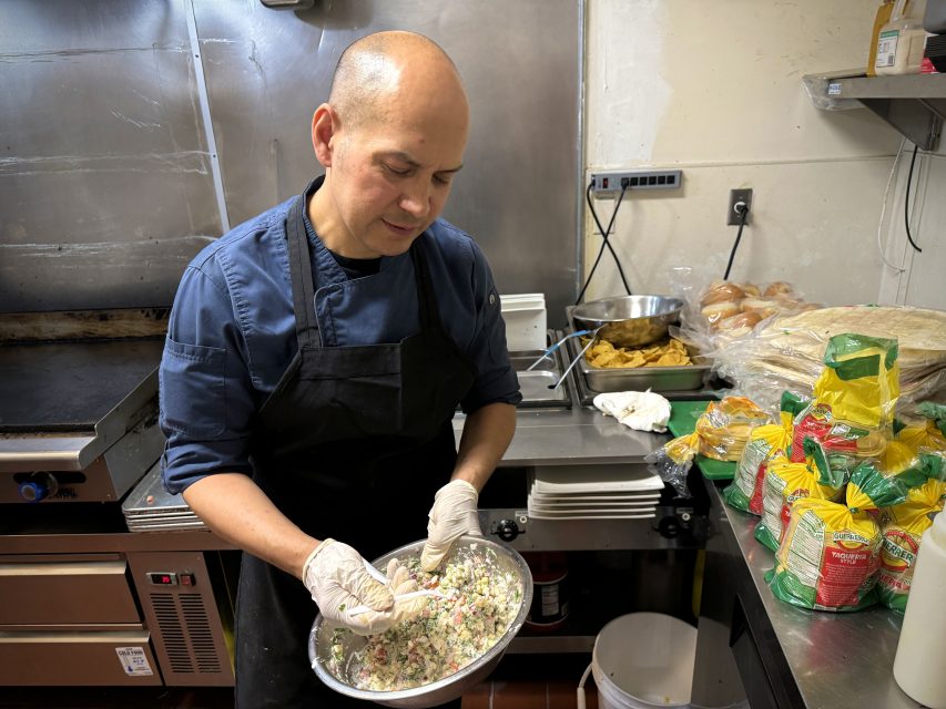 A person wearing gloves and a dark apron mixes food in a metal bowl in a commercial kitchen, with various cooking supplies and ingredients on the counter.
