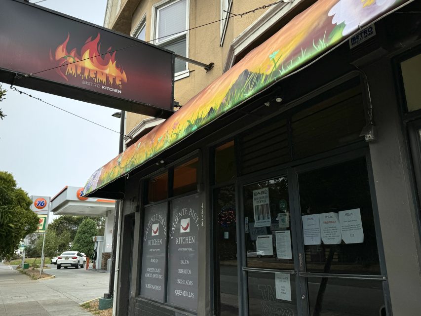 Street view of a restaurant called "Bistro Kitchen" with a colorful awning and a sign depicting flames. Multiple menus and notices are posted on the front windows. A 76 gas station is visible in the background.