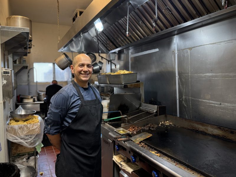 A person in a black apron smiles while standing next to a griddle in a commercial kitchen. Cooking utensils and ingredients are around the kitchen. Another person is visible in the background.