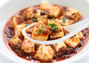 A spoon holding a piece of tofu in a spicy red sauce, with a dish of similarly prepared tofu in the background.