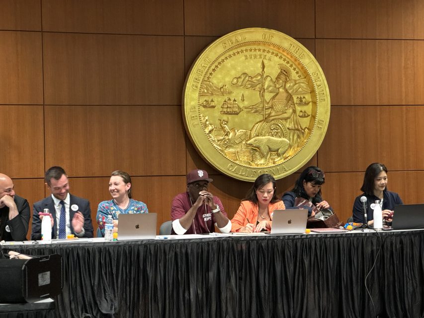 七人小組坐在一張長桌後,背後的木質鑲板牆上懸掛著一個大型圓形州印章徽章。部分成員正在使用筆記本電腦,其他人則在鼓掌或觀察。