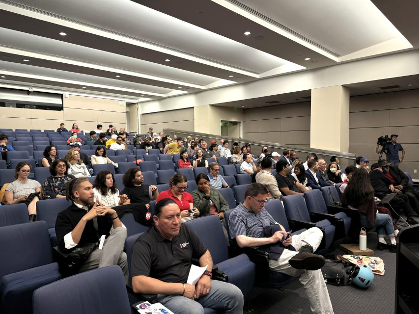 觀眾坐在一個演講廳內;有些人專注地觀看,其他人則在使用手機或交談。