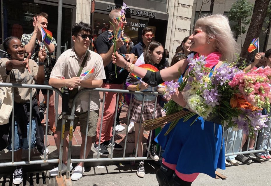 Person with a bouquet of flowers hands out floral stems to people behind a barrier during a pride event.