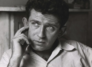 Black-and-white portrait of a man with short, dark hair, resting his head on his right hand while looking thoughtful. He is wearing a checkered button-up shirt.