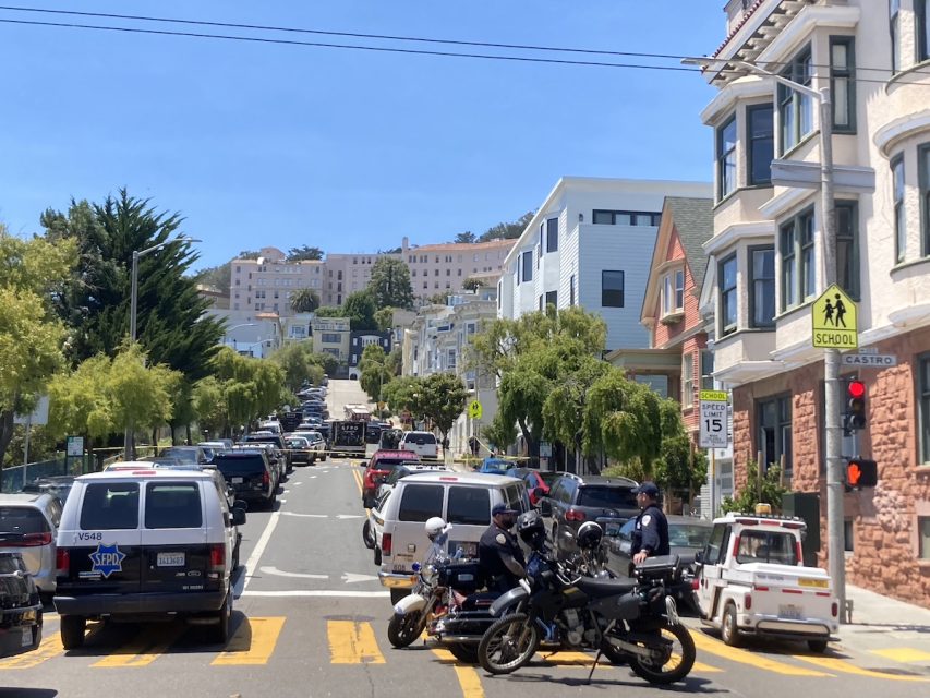 A city street busy with parked cars and traffic, police officers are stationed by their motorcycles near a crosswalk marked with school crossing signs and speed limit warnings.