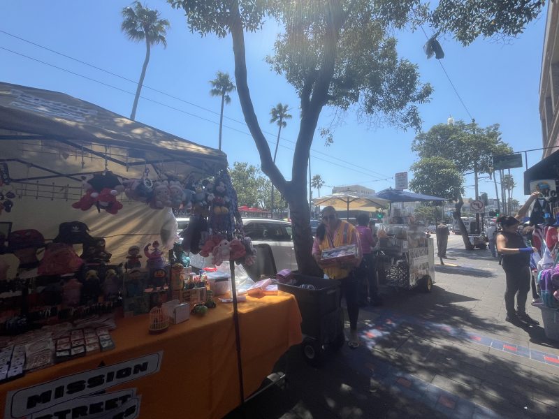 Outdoor street market with various stalls under a tree. One stall has toys on display, while another vendor is tending to a table. People are walking by under a sunny, clear sky.