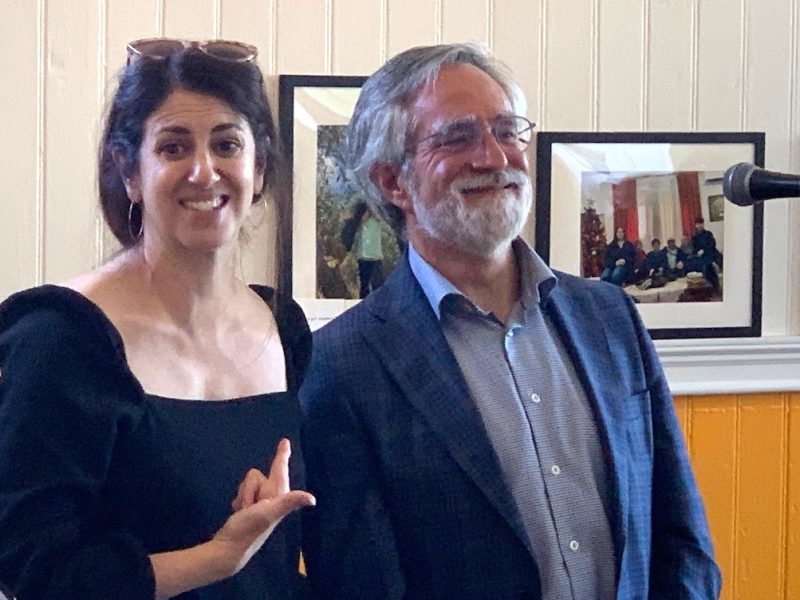 Two people, one smiling woman in a black dress pointing to a smiling man in a suit, standing indoors near a microphone with photo frames on the wall behind them.