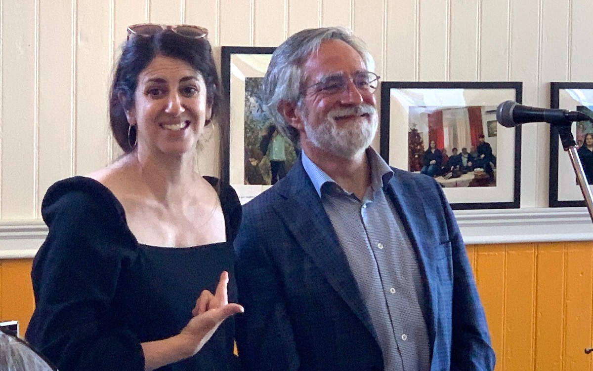 Two people, one smiling woman in a black dress pointing to a smiling man in a suit, standing indoors near a microphone with photo frames on the wall behind them.