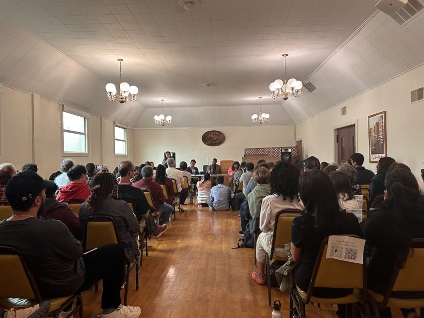 A group of people sits in a well-lit room facing a panel of speakers. The room has wooden floors, chandeliers, and cream-colored walls with windows on one side.