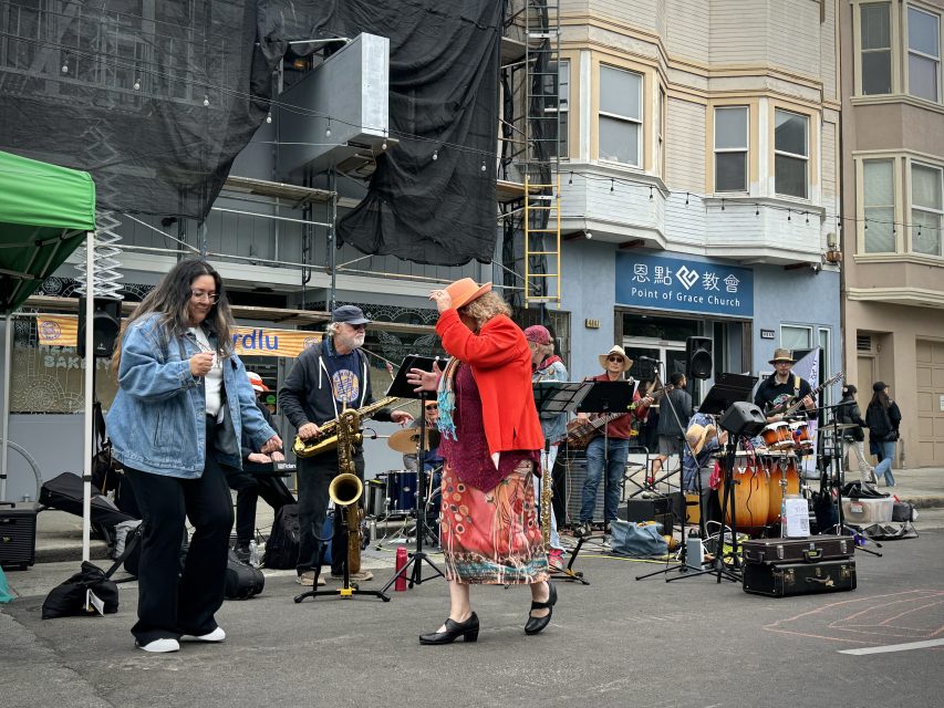 A band performs on a street next to a building under construction, while two people dance in the foreground. Musical instruments include saxophone, drums, and keyboard.