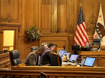 Four individuals sit at a wooden desk with multiple computer monitors in a room with ornate wood paneling, an American flag, and a California state flag.