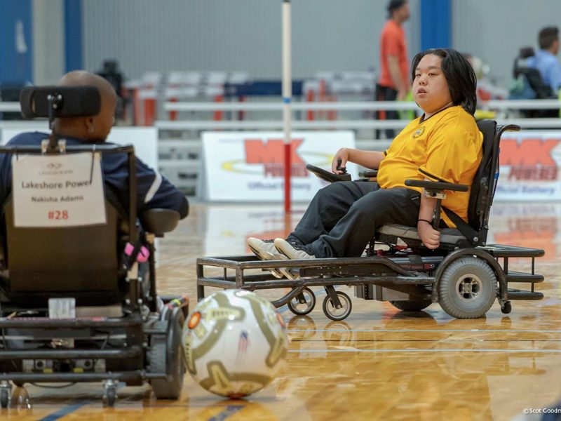 Two individuals in power wheelchairs playing power soccer indoors. One player in a yellow jersey looks towards the ball near them, while the other player in a black jersey is seen from behind.