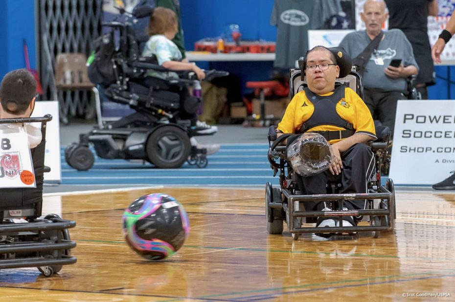 A man in a powered wheelchair wearing a yellow and black shirt plays power soccer on a hardwood court, with a colorful ball in motion nearby. Other participants and spectators are visible in the background.