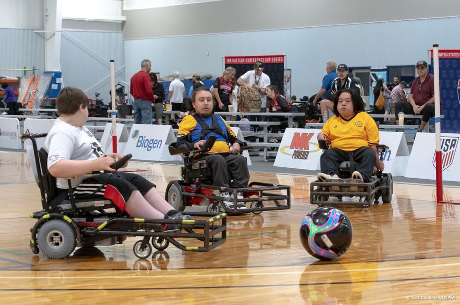Three athletes in power wheelchairs are on an indoor court with a colorful soccer ball. Spectators and officials can be seen in the background.