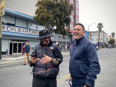 Two men stand in a city street, smiling and engaged in conversation. One is looking at a smartphone, and the other is laughing. A childcare center is visible in the background.