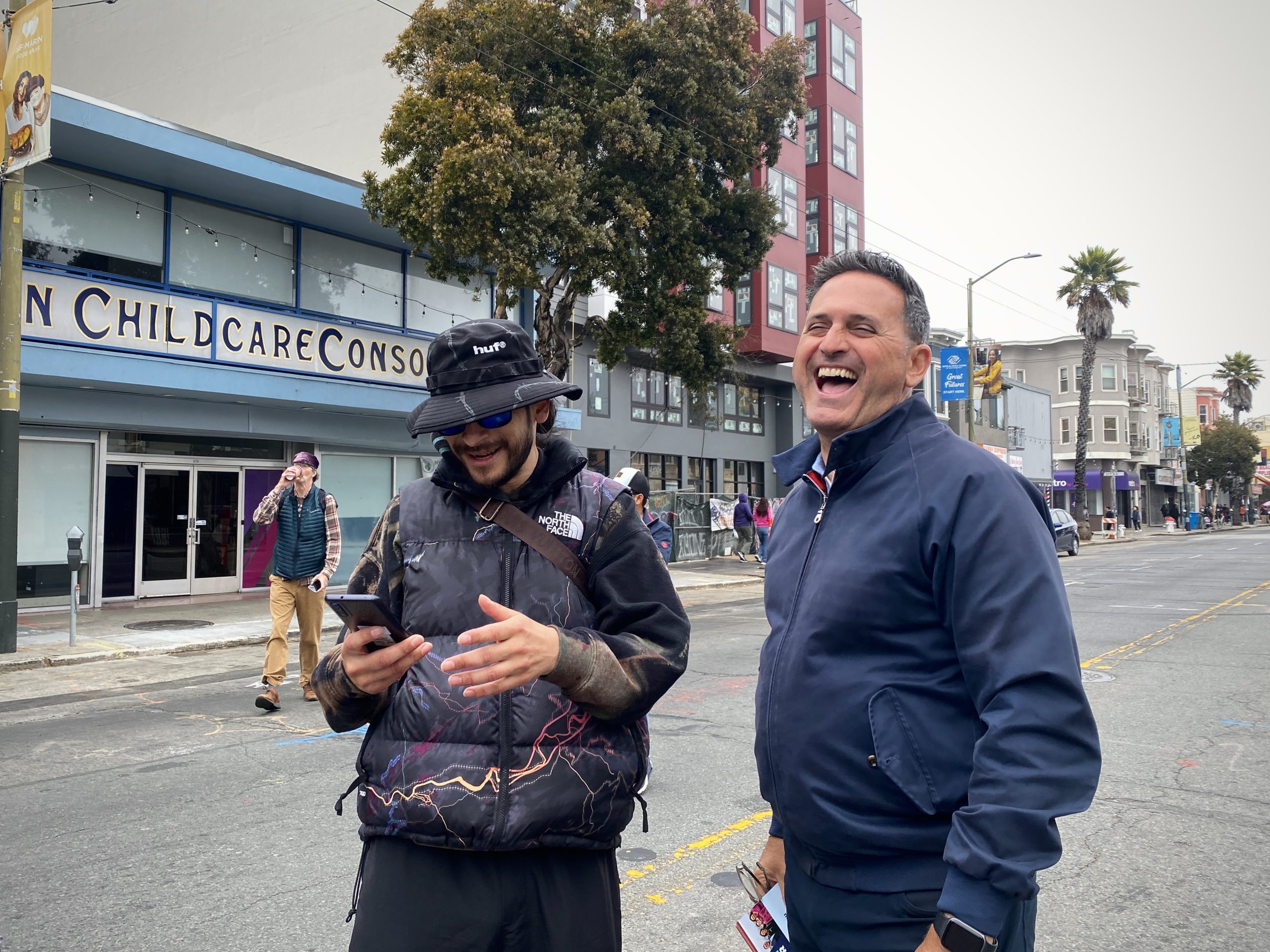 Two men stand in a city street, smiling and engaged in conversation. One is looking at a smartphone, and the other is laughing. A childcare center is visible in the background.
