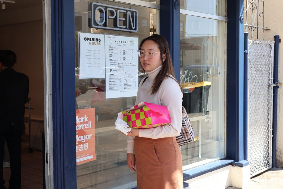 A person stands outside a storefront with an "OPEN" sign, holding a pink and green package and a small item. A menu and opening hours are posted on the glass door.