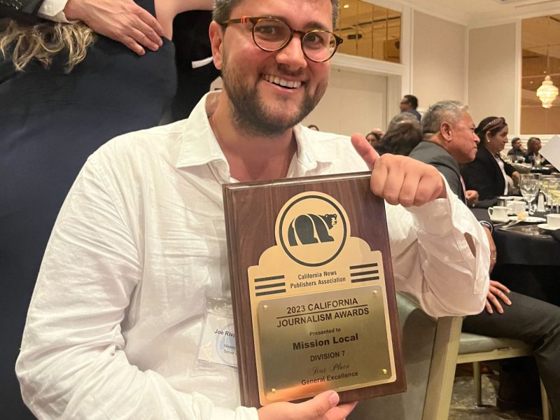 A man in a white shirt and pink pants holds a plaque for the 2023 California Journalism Awards at a formal event. Other attendees can be seen in the background, including two people hugging.
