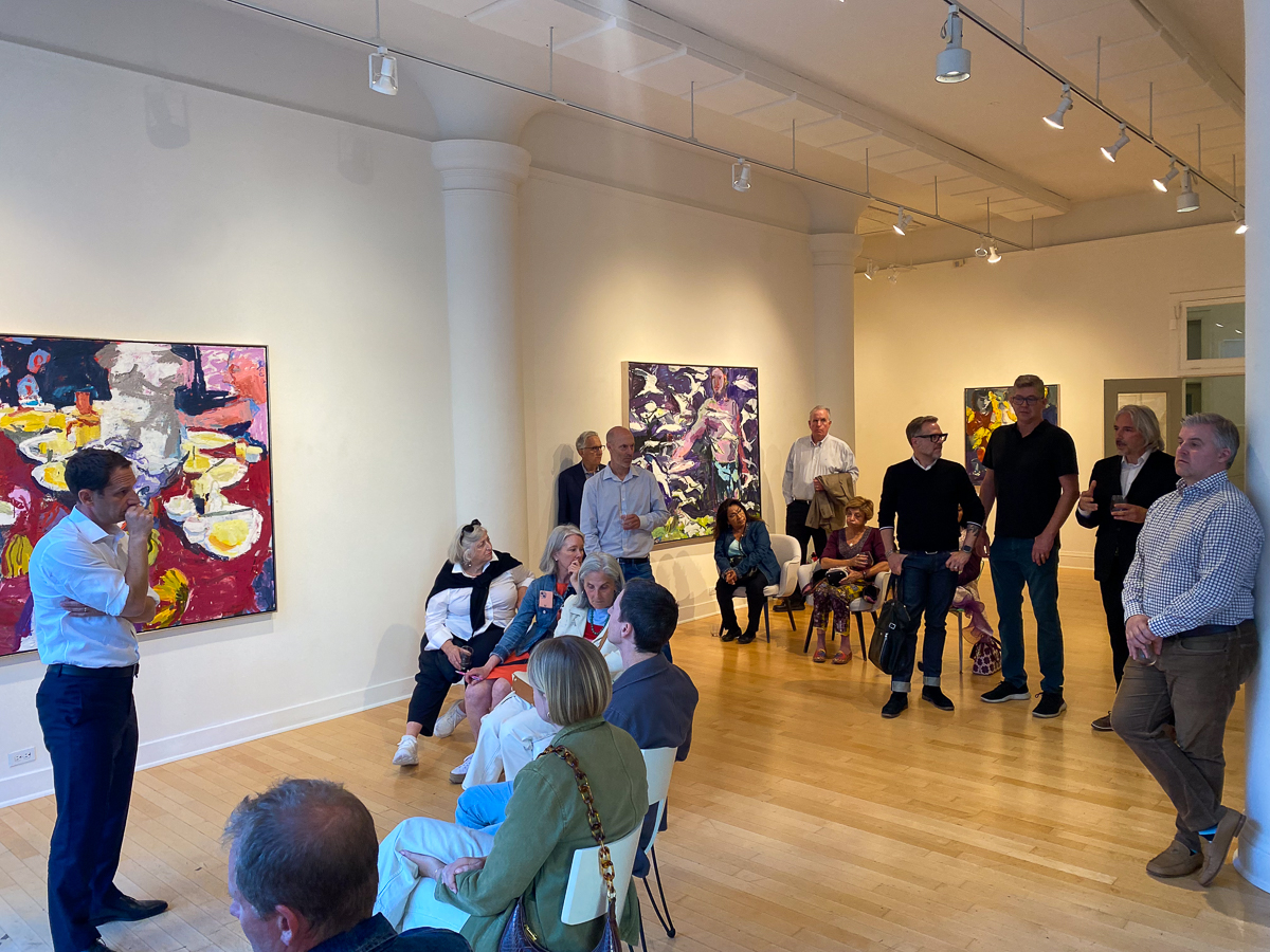 A group of people attend an art gallery presentation, standing and sitting while listening to a speaker. Paintings are displayed on the walls.