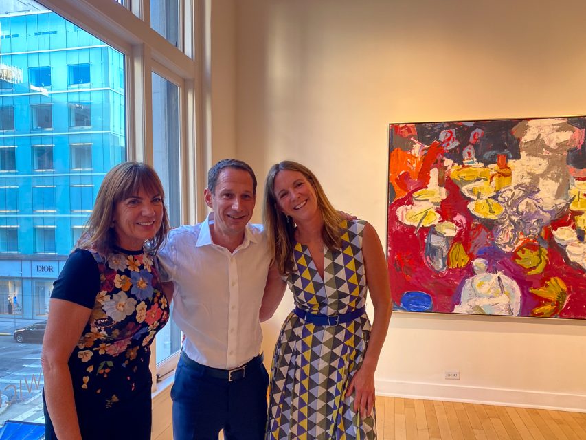 Three people smiling and standing close together in an art gallery, with abstract paintings visible on the walls and large windows showing a cityscape in the background.
