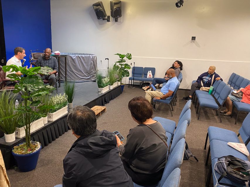 A small group of people are seated on blue chairs in a room, attentively listening to two men speaking on a small stage with plants in the foreground.