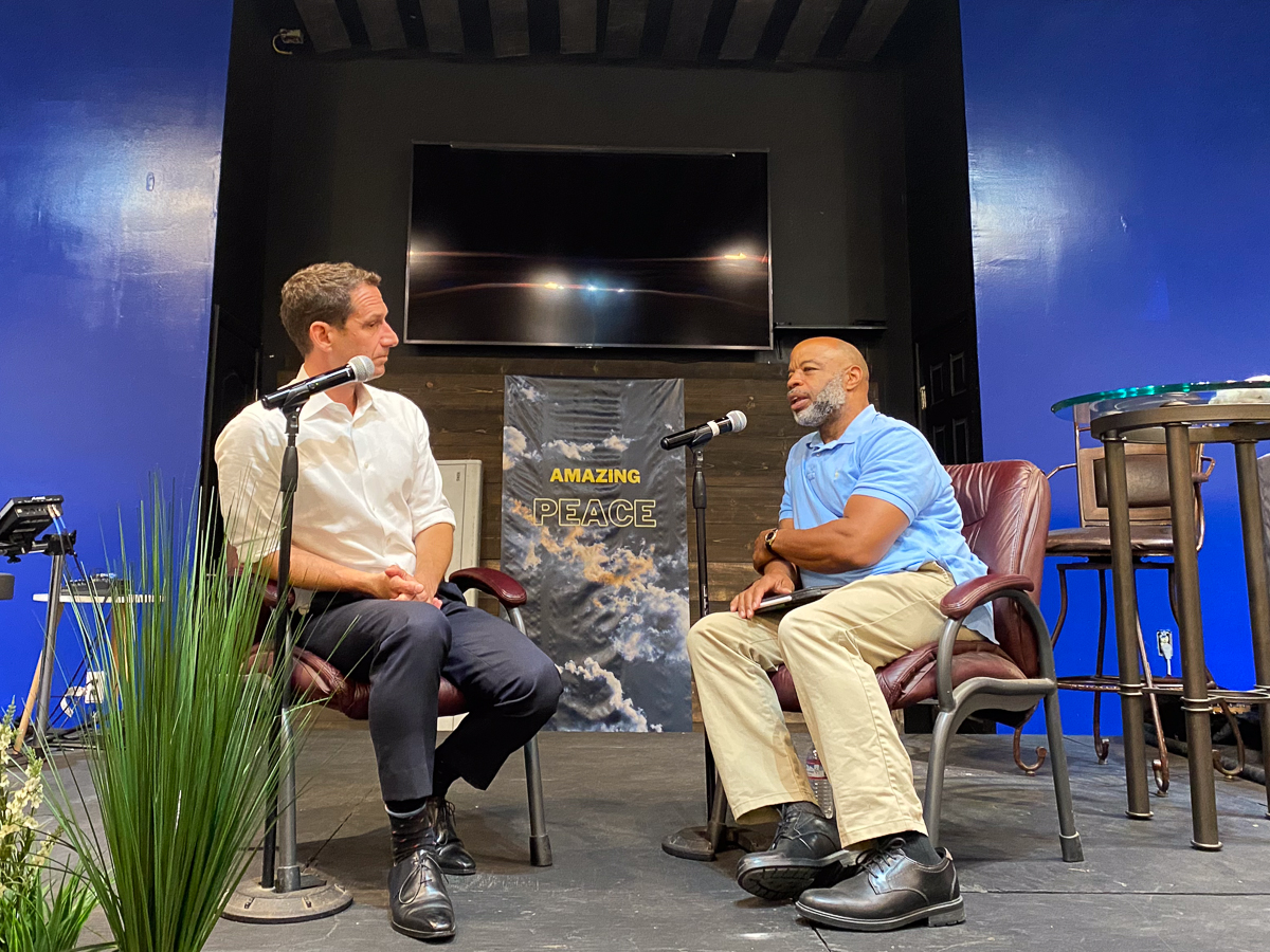 Two men sit on a stage with microphones in a discussion setting. Behind them is a banner that reads "AMAZING PEACE." The background is blue with a TV screen above them.