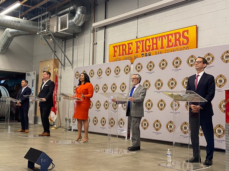 Five individuals in formal attire stand behind podiums on a stage in front of a "Fire Fighters Local Union No. 798 San Francisco" sign.