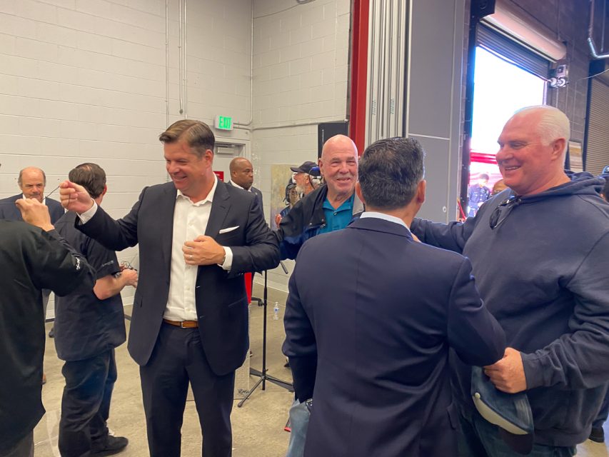 A group of men in casual and business attire engage in conversation and greet each other indoors, with a partially open garage door in the background.