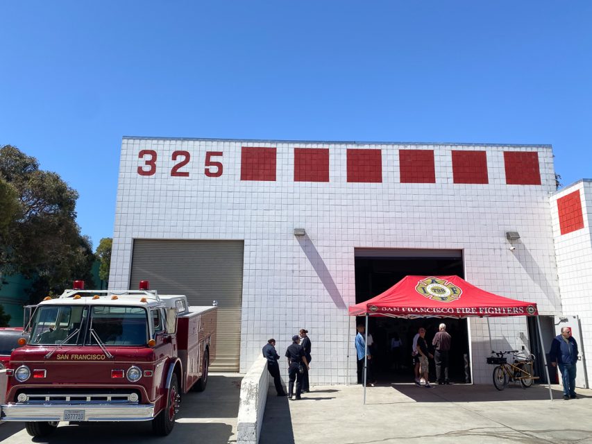 A classic red fire truck is parked in front of a white brick fire station numbered 325. Several people stand near an open garage and a red tent labeled "San Francisco Fire Fighters.