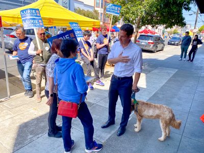 A man in a white shirt engages in conversation with two people on a sidewalk while holding a dog on a leash. Campaign supporters holding signs and wearing shirts gather under yellow tents in the background.