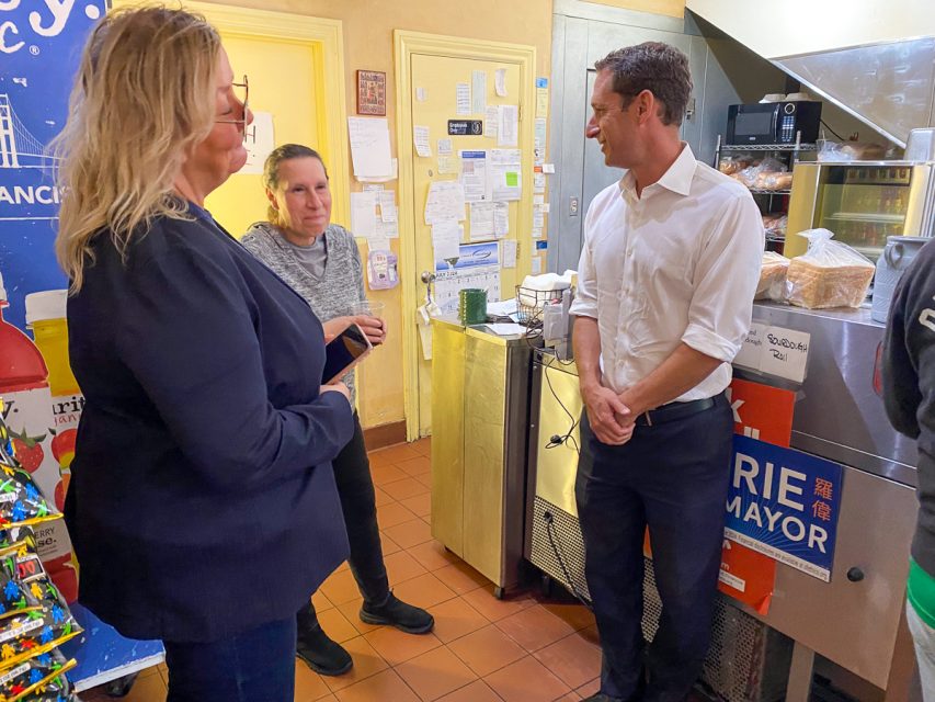 Three people are talking in a room with a counter, refrigerator, and various posters and notes on the walls. One person is holding a book. The room appears to be a casual, informal setting.