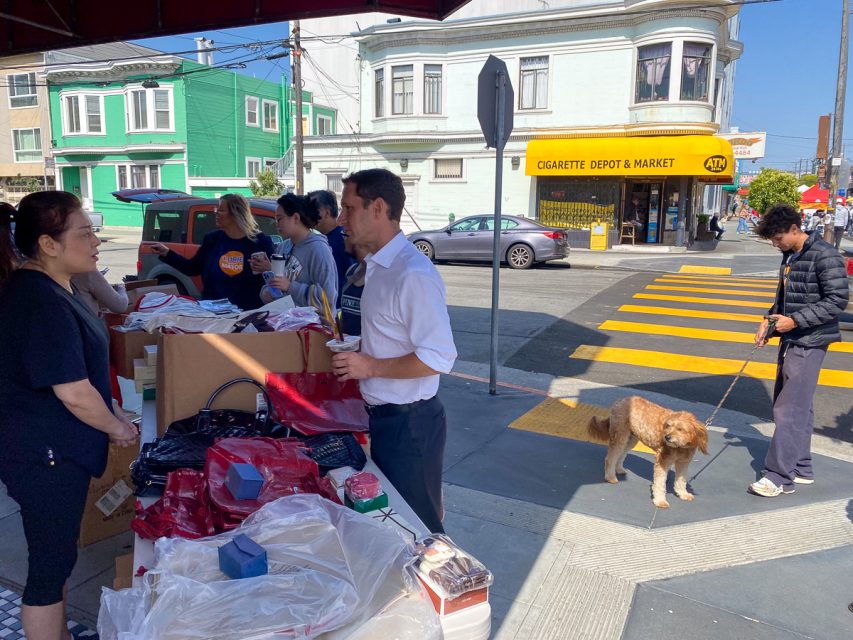 People interacting at an outdoor market stall on a sunny day in a city. One person is holding a dog on a leash while others are examining items on a table. A store sign reads "Cigarette Depot & Market".