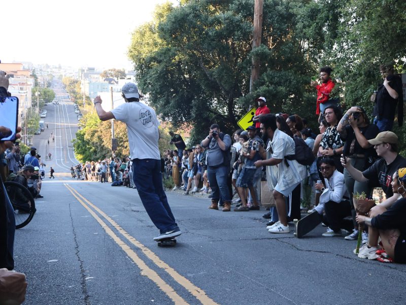 A skateboarder rides down a sloped street as a large crowd gathered on both sides watches and takes photos. Trees and buildings are visible in the background.