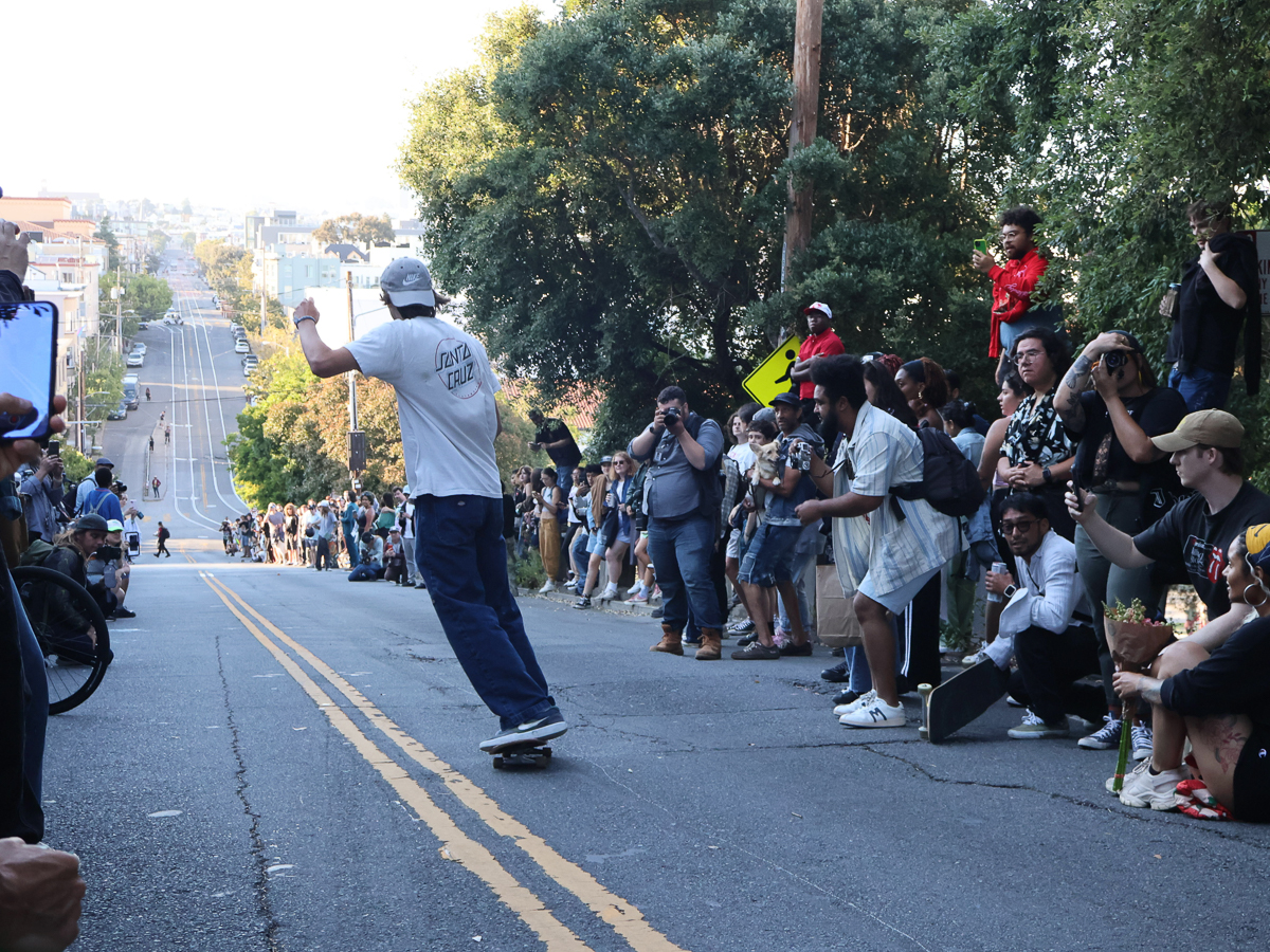 A skateboarder rides down a sloped street as a large crowd gathered on both sides watches and takes photos. Trees and buildings are visible in the background.