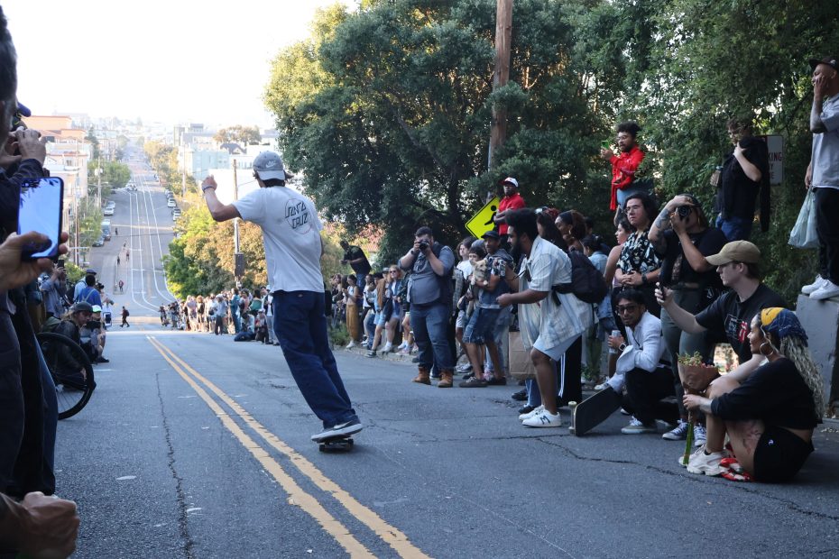 A skateboarder rides down a hilly street lined with cheering spectators, some taking photos, while others watch from the sidelines.