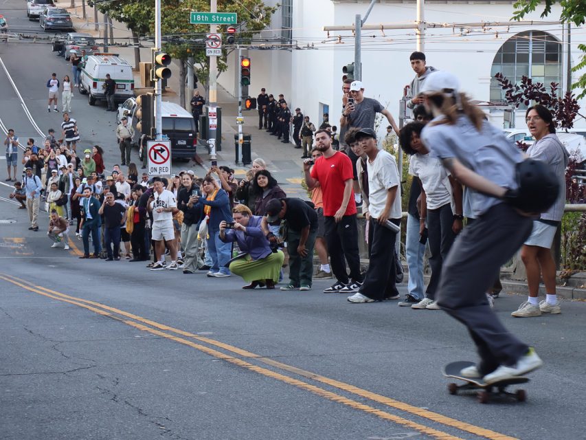 A skateboarder rides down a hill as a crowd of onlookers stands and photographs along the street. The scene includes street signs, parked cars, and buildings in the background.