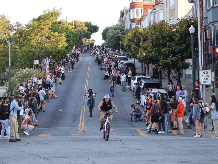 A crowd gathers along a sloped urban street, watching cyclists ride towards them. Tall row houses and trees line the sidewalk. The mood appears lively and engaged.