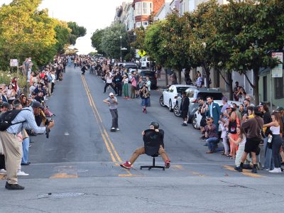 People line both sides of the street as skateboarders and a person on an office chair roll downhill in an urban setting. Photographers capture the scene.