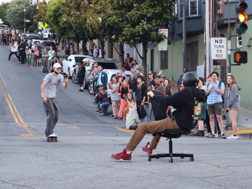 A person in an office chair and another on a skateboard race down a street as a large crowd cheers from the sidewalks. A "No Turn On Red" sign and parked cars are visible in the background.
