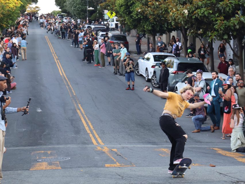 A skateboarder performs a trick on a downhill street while spectators line both sides of the road, capturing the moment with their phones.