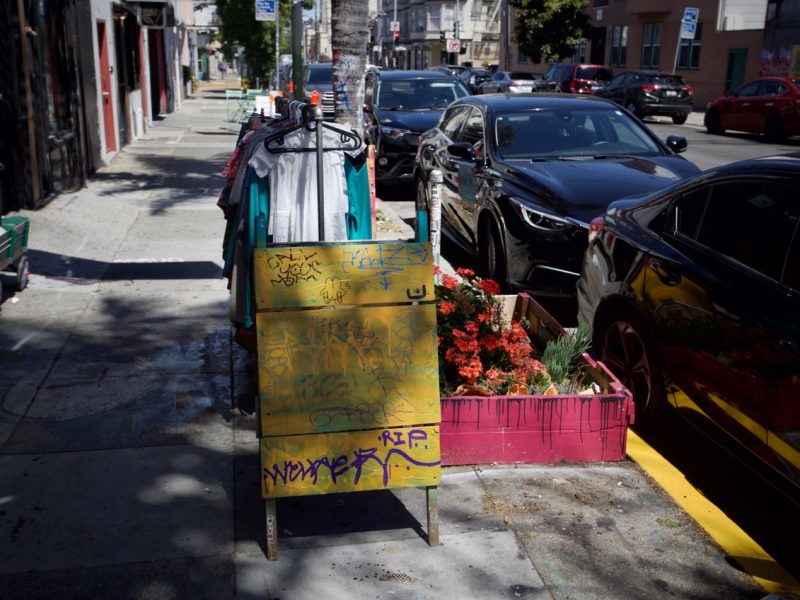 A city sidewalk scene showing a graffiti-covered yellow sidewalk sign, a row of parked cars, potted red flowers, and clothes hanging on racks under partial sunlight and shade.